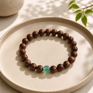May include: A circular bracelet composed of numerous dark brown, polished wooden beads with a visible grain pattern, featuring a single smooth, translucent light green bead positioned as a focal point within the continuous strand.