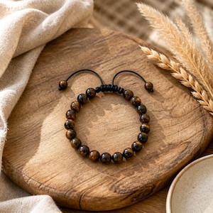 Könnte beinhalten: Ein Perlenarmband mit polierten, runden Tigerauge-Edelsteinen in goldbraunen und schwarzen Farbtönen. Die Perlen sind auf eine schwarze Kordel mit einem verstellbaren Schiebeknotenverschluss aufgezogen, der an den Enden mit zwei kleinen Perlen versehen ist.