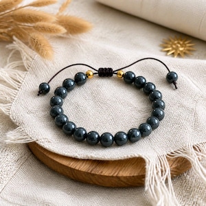 May include: A dark grey beaded bracelet featuring smooth, round, polished stones arranged in a circular pattern. The bracelet includes two small, shiny gold-toned metal beads near the adjustable dark brown cord closure with two dangling end beads.