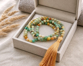 Peut inclure: Un collier de perles avec un pompon, présenté dans un coffret clair ouvert. Le collier présente une variété de perles aux tons terreux, notamment vertes, jaunes et marron. Le pompon est de couleur or chaud, ajoutant une touche d'élégance.