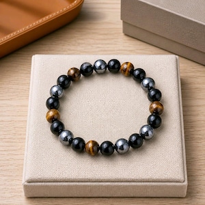 Peut inclure: Un bracelet composé de perles rondes et lisses disposées en alternance, présentant des tons noirs, gris métallisé et brun doré façon œil-de-tigre. Les perles sont assemblées en cercle, offrant une finition polie et des textures minérales variées.