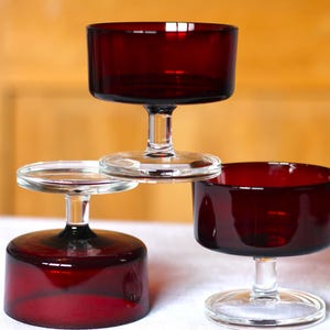 Può includere: Tre bicchieri vintage in vetro rosso rubino per dessert o cocktail. Ogni bicchiere presenta un gambo corto e una coppa ampia e poco profonda. I bicchieri sono disposti in una composizione impilata, che mette in risalto il loro design e colore.