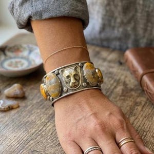 Könnte beinhalten: Silbernes Armreif-Armband mit einem zentralen Katzenkopf-Design und ovalen gelb-grauen Steinen. Das Armband wird am Handgelenk getragen und zeigt die detaillierte Gestaltung und Handwerkskunst des Schmucks.
