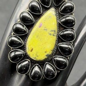 Może przedstawiać: Srebrny pierścionek z owalnym, żółto-zielonym kamieniem pośrodku, otoczonym czarnymi kamieniami w kształcie łezki. Pierścionek jest prezentowany na czarnej powierzchni.