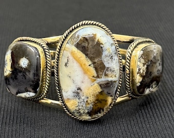 Peut inclure: Bracelet en métal doré orné de trois pierres polies. La pierre centrale est ovale, flanquée de deux pierres carrées. Les pierres présentent des motifs blancs, bruns et jaunes, créant un aspect naturel unique.