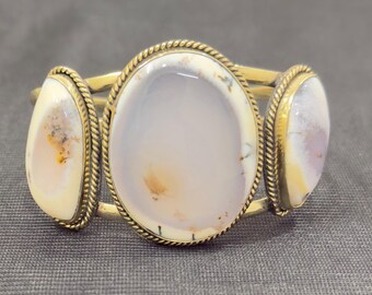 Peut inclure: Un bracelet manchette doré avec trois grandes pierres ovales. Les pierres ont une base blanc crème avec des inclusions brunes et violettes. Chaque pierre est encadrée d'une bordure torsadée en forme de corde.