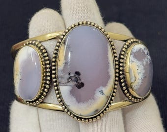 Peut inclure: Un bracelet manchette en métal doré avec trois pierres ovales de couleur violet clair aux motifs uniques. Les pierres sont serties dans un bord perlé. Ce bracelet est un bijou.