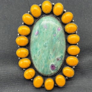 Może przedstawiać: Owalny pierścionek z dużym, jasnozielonym kamieniem z różowymi wtrąceniami, otoczonym obramowaniem z pomarańczowych kamieni. Pierścionek ma srebrzystą metalową oprawę, a tło jest ciemnoszare.