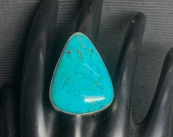 May include: A large, triangular turquoise ring with a silver band. The turquoise stone has a vibrant blue-green color with brown veining. The ring is set on a black surface.