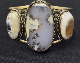 Peut inclure: Bracelet manchette argenté orné de trois pierres ovales. La pierre centrale est grise clair avec des inclusions noires et jaunes. Les pierres latérales présentent des motifs crème et marron. Les pierres sont serties dans un cadre métallique décoratif.