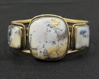Peut inclure: Un bracelet manchette doré avec trois pierres rectangulaires et carrées. Les pierres sont blanches avec des inclusions noires et beiges. Le bracelet est posé sur un fond sombre.