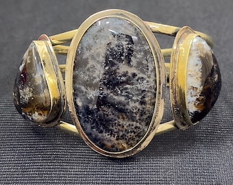 Peut inclure: Un bracelet manchette en métal doré avec trois pierres naturelles. La pierre ovale centrale est principalement noire et grise, tandis que les pierres latérales sont en forme de goutte avec des motifs bruns et blancs. Le bracelet a une finition polie.