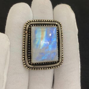 May include: A rectangular silver ring with a large, iridescent moonstone. The moonstone has a blue and white glow. The ring has a beaded border and a square frame around the stone.