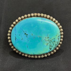 May include: Oval turquoise gemstone set in a silver-toned metal frame. The turquoise stone is a vibrant blue-green color with dark veining. The frame has a detailed, beaded edge, creating a decorative border. The jewelry piece is set against a dark background.