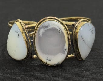 Peut inclure: Bracelet doré orné de trois pierres blanches avec des inclusions grises et noires. Les pierres sont de forme ovale et en goutte. Le bracelet présente un design simple et élégant, adapté au quotidien ou aux occasions spéciales.