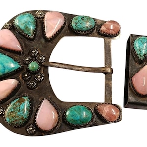 May include: A silver-toned belt buckle and keeper set. The buckle is adorned with turquoise and pink gemstones in a decorative pattern. The keeper has matching gemstones. The buckle is approximately 3 inches wide.