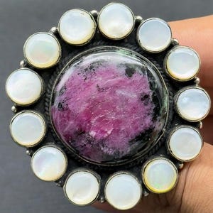 Może przedstawiać: Srebrny pierścionek z dużym, okrągłym, fioletowo-różowym kamieniem pośrodku, otoczonym aureolą z białych, okrągłych kamieni z masy perłowej. Pierścionek ma szczegółową, teksturowaną oprawę.