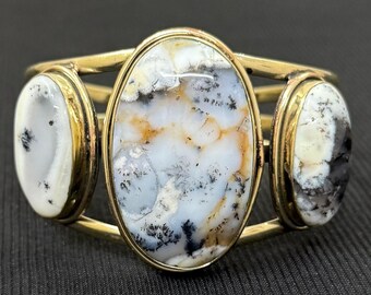 Peut inclure: Un bracelet manchette en métal doré avec trois pierres ovales. Les pierres présentent des motifs blancs, noirs et beiges. Le bracelet a un design simple et élégant, avec les pierres serties dans des chatons individuels.