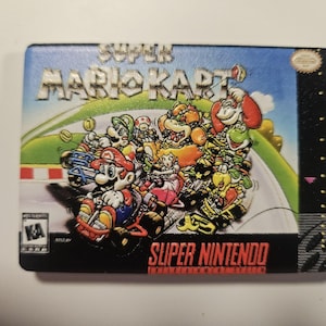 Puede incluir: Un cartucho de videojuego Super Mario Kart. El cartucho presenta una colorida ilustraci&oacute;n de Mario y otros personajes corriendo en una pista. Las palabras "SUPER MARIO KART" y "SUPER NINTENDO" se muestran de forma destacada.
