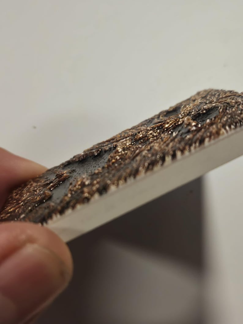 Puede incluir: Un objeto rectangular con una superficie texturizada. La superficie superior tiene una textura oscura y rugosa con tonos bronce y negro. Los lados son de color claro y liso. El objeto se sostiene en &aacute;ngulo.