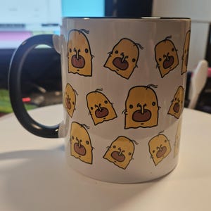 Peut inclure: Mug en céramique blanche avec une anse et un bord noirs. Le mug est orné d'un motif répété de visages de dessins animés jaunes avec des détails marron. Le mug est posé sur une surface blanche.