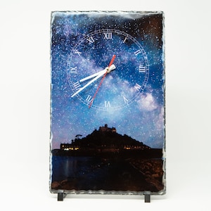 May include: A slate clock with a black metal stand featuring a night sky image with a white clock face and Roman numerals. The image also includes a silhouette of a castle on a small island.