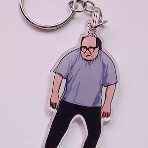 Könnte beinhalten: Schlüsselanhänger mit einer Cartoon-Illustration eines Mannes mit Brille, grauem T-Shirt und schwarzer Hose. Der Schlüsselanhänger hat einen silbernen Schlüsselring und Verschluss.