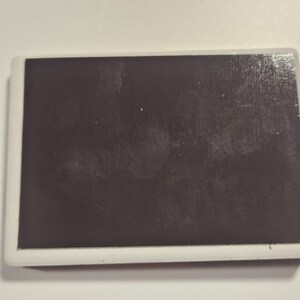Puede incluir: Im&aacute;n rectangular con una superficie marr&oacute;n oscuro y un borde blanco. El im&aacute;n est&aacute; sobre un fondo blanco. La superficie del im&aacute;n parece tener algunas imperfecciones menores.
