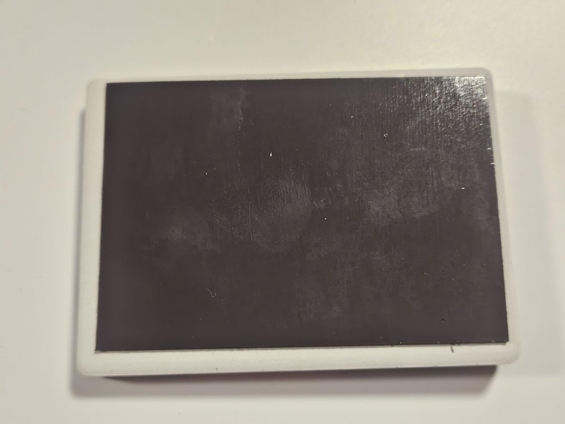 Puede incluir: Im&aacute;n rectangular con una superficie marr&oacute;n oscuro y un borde blanco. El im&aacute;n est&aacute; sobre un fondo blanco. La superficie del im&aacute;n parece tener algunas imperfecciones menores.