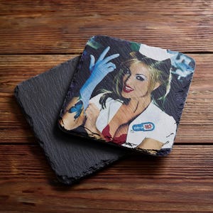 May include: Two square slate coasters with rough, natural edges. One coaster is plain dark gray, while the other features a printed image of a person in a white nurse uniform and cap, wearing blue gloves, with a Blink-182 logo on the chest area.