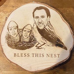 Puede incluir: Una rodaja de madera con un diseño grabado con láser que presenta un pájaro con la cara de Nicolas Cage y tres caras de Nicolas Cage en un nido. El texto "BLESS THIS NEST" está grabado debajo.