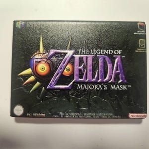 Op de afbeelding: Gamebox van The Legend of Zelda: Majora's Mask. De doos is zwart met de speltitel in paars en het iconische masker met gele ogen. Het Nintendo-logo staat rechtsonder.