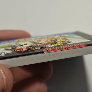 Puede incluir: Un cartucho de videojuego de Super Nintendo. El cartucho del juego tiene una imagen colorida de coches de carreras en la parte superior. El t&iacute;tulo del juego "Super Nintendo" est&aacute; impreso en letras rojas en el lateral del cartucho.