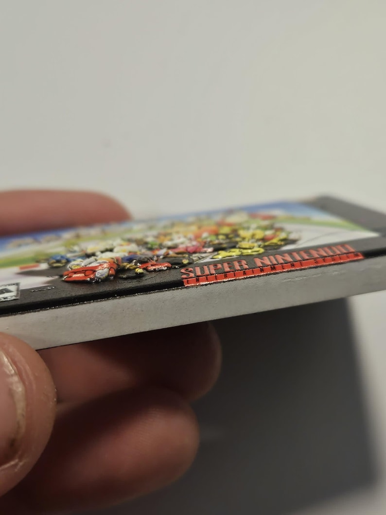 Puede incluir: Un cartucho de videojuego de Super Nintendo. El cartucho del juego tiene una imagen colorida de coches de carreras en la parte superior. El t&iacute;tulo del juego "Super Nintendo" est&aacute; impreso en letras rojas en el lateral del cartucho.