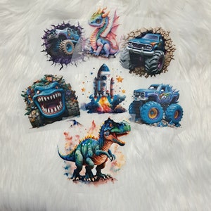 Könnte beinhalten: Sechs Bügelbilder mit verschiedenen Designs, darunter ein blauer Monstertruck, ein blauer Dinosaurier, ein blauer Drache, eine blaue Rakete, ein blauer Monstertruck und ein blauer Monstertruck. Die Bügelbilder sind alle auf weißem Hintergrund.