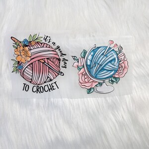Könnte beinhalten: Zwei Bügelbilder mit einem rosa und blauen Wollknäuel mit Stricknadeln und Blumen. Der Text auf einem Bügelbild lautet "Es ist ein guter Tag zum Häkeln".