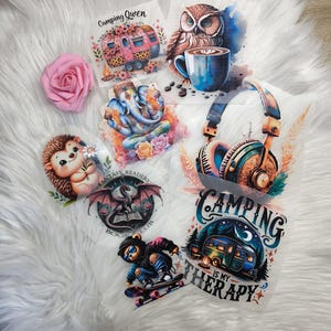 Könnte beinhalten: Ein Set mit sechs Bügelbildern mit bunten Illustrationen. Die Designs beinhalten eine Campingkönigin mit einem Wohnwagen, eine Eule mit einer Kaffeetasse, einen meditierenden Elefanten, einen Igel mit Blumen, einen Drachen mit einem Buch und einen Bären auf einem Skateboard mit dem Text "Camping is my therapy".