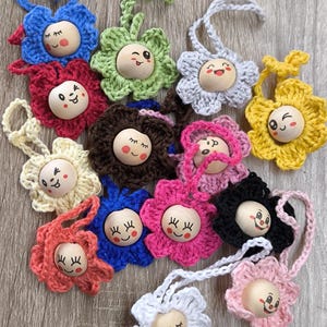 Könnte beinhalten: Eine Sammlung bunter, gehäkelter Blumenornamente mit Gesichtern aus Holzperlen. Die Blumen sind in verschiedenen Farben erhältlich, darunter Blau, Rosa, Gelb und Weiß. Jede Blume hat ein lächelndes Gesicht und eine Schlaufe zum Aufhängen.
