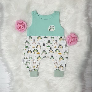 May include: A light blue and white baby romper with a panda bear and rainbow print. The romper has a sleeveless top and a pocket on the front.