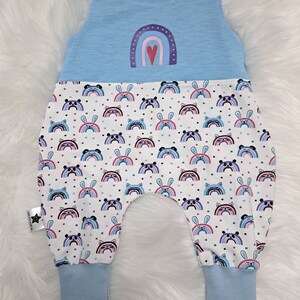 May include: Light blue baby overalls with a rainbow and heart design on the top and a white bottom with a repeating pattern of rainbows, bunnies, and panda faces.