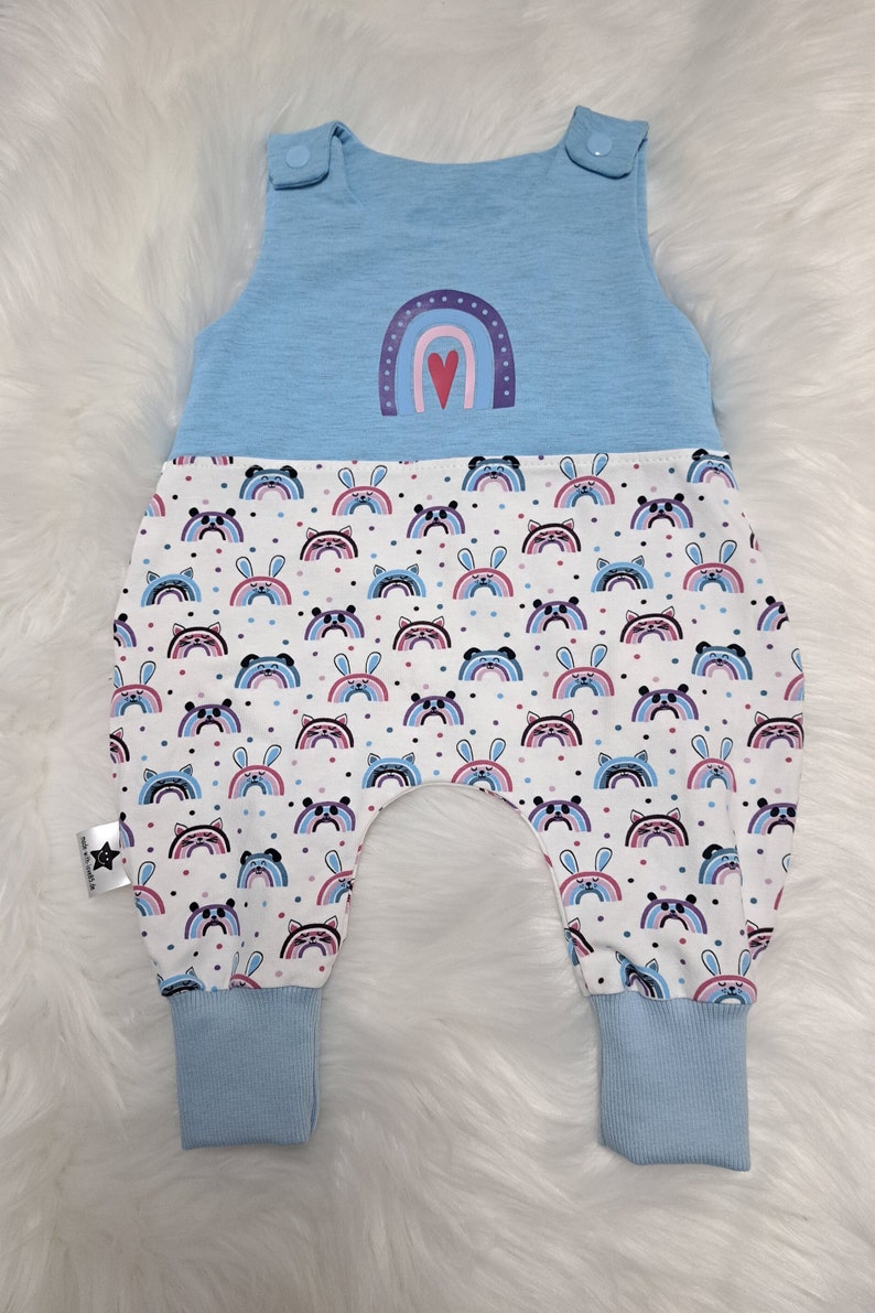 May include: Light blue baby overalls with a rainbow and heart design on the top and a white bottom with a repeating pattern of rainbows, bunnies, and panda faces.