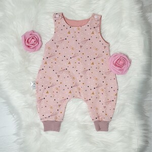 May include: Pink baby overall with a star and shooting star print. The overall has a pink and white color scheme with a subtle gold glitter effect.