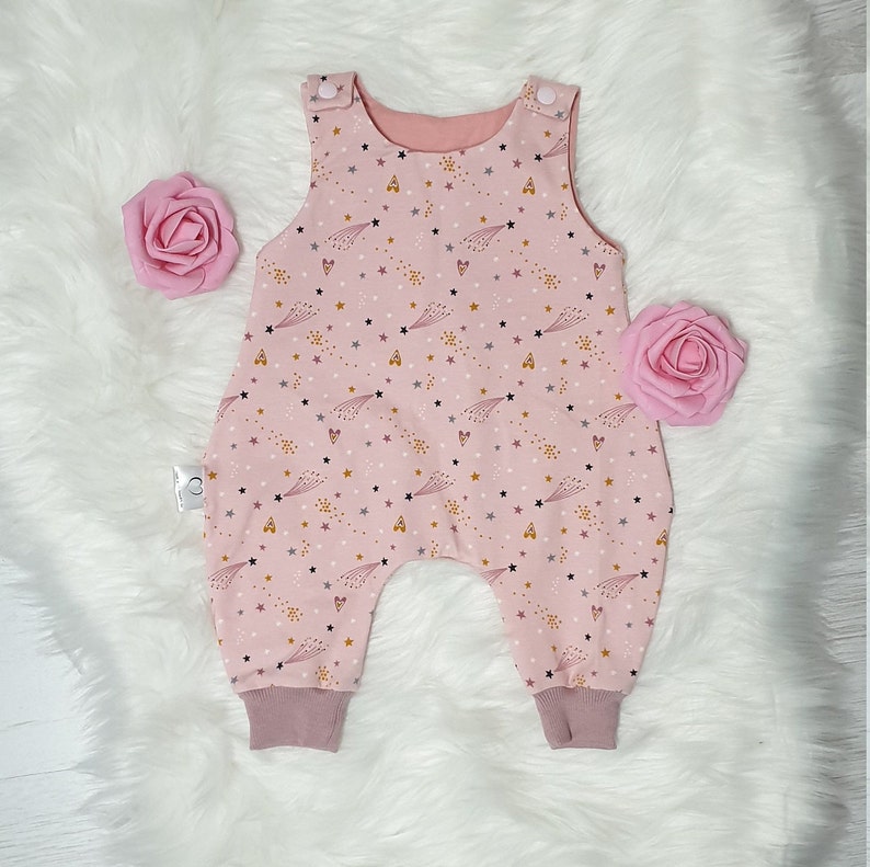 May include: Pink baby overall with a star and shooting star print. The overall has a pink and white color scheme with a subtle gold glitter effect.