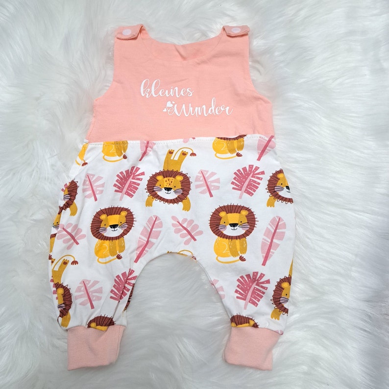 May include: A pink and white baby onesie with a lion print. The onesie has a pink top with the text "kleines Wunder" in white.