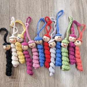 Könnte beinhalten: Eine Sammlung handgefertigter Häkelpuppen mit Holzköpfen und lächelnden Gesichtern. Jede Puppe hat ein anderes Farbschema, darunter Schwarz, Weiß, Gelb, Rosa, Blau, Rot, Grau, Grün und Koralle. Die Puppen haben Schlaufenschnüre zum Aufhängen.