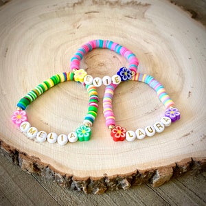 May include: Three colorful beaded bracelets with flower charms and names spelled out in white letters. The bracelets are Amelia, Evie, and Laura.