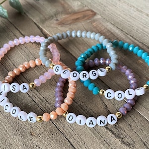 Peut inclure: Quatre bracelets en perles avec des perles de différentes couleurs et des accents dorés. Chaque bracelet porte un nom : Luisa, Nicole, George et Diana.