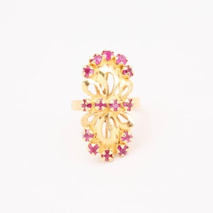 May include: A gold-colored ring with a floral design, adorned with multiple small, round, red gemstones. The ring's intricate filigree work and the vibrant red stones create a striking contrast against the gold setting. The ring is set against a white background.