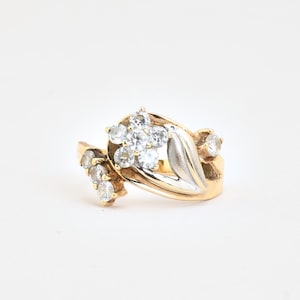 Puede incluir: Un anillo de oro con un grupo central en forma de flor de cinco piedras redondas transparentes, acentuado por un diseño de hoja curva en tono plateado y dos piedras redondas transparentes pequeñas adicionales en el lateral, sobre una banda de oro amarillo.
