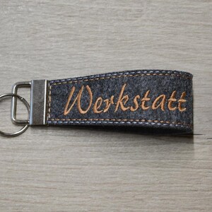 Könnte beinhalten: Ein grauer Filzschlüsselanhänger mit einem silbernen Metallring und einem blauen gestickten Rand. Das Wort "Werkstatt" ist in orangefarbenem Garn gestickt.